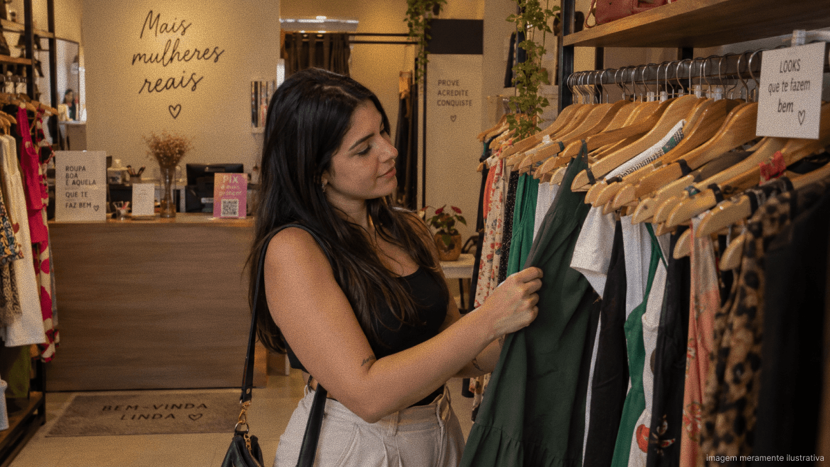 Mulher escolhendo uma peça de roupa em uma loja de moda feminina local, com araras de roupas e ambiente acolhedor.